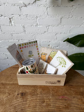 A wooden gift box filled with gift items such as an insulated mug, watercolor brushes and a watercolor workbook, crystals, a tee shirt, and a birthday calendar, sit atop a wooden table in front of a white brick background. 