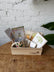 A wooden gift box filled with gift items such as an insulated mug, watercolor brushes and a watercolor workbook, crystals, a tee shirt, and a birthday calendar, sit atop a wooden table in front of a white brick background. 