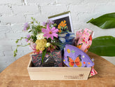 A wooden gift box filled with a floral arrangement, playing cards, a zippered pouch, socks, a candle, and a botanical print card, sitting on top of a wooden table in front of a white brick background. 