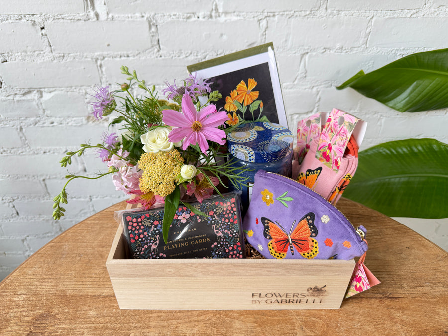 A wooden gift box filled with a floral arrangement, playing cards, a zippered pouch, socks, a candle, and a botanical print card, sitting on top of a wooden table in front of a white brick background. 