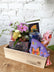 A side shot of a wooden giftbox filled with a floral arrangement and other gift items, sitting on top of a wooden table in front of a white brick background.