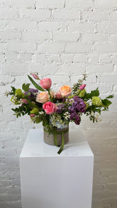 A vibrant floral arrangement featuring pink and peach roses, purple lilac, blue hydrangeas, and green viburnum, presented in a burlap-wrapped vase with a green ribbon, sitting on a white pedestal against a white brick wall.