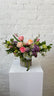 A vibrant floral arrangement featuring pink and peach roses, purple lilac, blue hydrangeas, and green viburnum, presented in a burlap-wrapped vase with a green ribbon, sitting on a white pedestal against a white brick wall.
