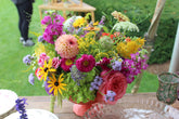 A vibrant, multicolor floral arrangement with pink, yellow, purple and green flowers, in a pink compote on an outdoor table.