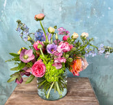 A vibrant, multicolored floral arrangement featuring pink, purple, orange, and blue flowers with green foliage, presented in a clear glass vase on a rustic wooden table against a textured light blue wall.