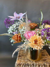 A vibrant floral arrangement made with a base of babies breath, in a gold leaf vase, displayed on a wicker stand in front of a grey background.