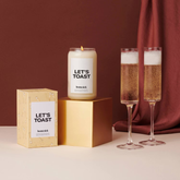 Two champagne glasses sit next to a candle and the candle's box, in front of a brick red background. 
