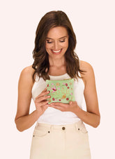Woman holding a mint-green mini velvet pouch with embroidered flowers, a bird, and a bee, with the word "LOVE" in the center. The woman is wearing a white shirt and beige pants, standing against a white background.