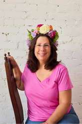 A woman in a hot pink shirt wearing a flower crown sits sideways on a dark wooden chair in front of a white brick background.