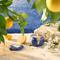 A blue tin candle sitting in the sand with lemons and white flowers framing it, and bright blue water in the background.