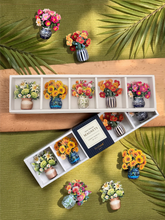 A stylized photo of a green background with palm leaves and a wooden pattern across the middle. On top of the flat-lay are two boxes with five mini paper bouquets, each with different flowers, and one of each bouquet scattered across the rest of the photo.
