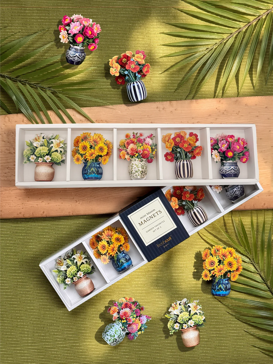 A stylized photo of a green background with palm leaves and a wooden pattern across the middle. On top of the flat-lay are two boxes with five mini paper bouquets, each with different flowers, and one of each bouquet scattered across the rest of the photo.