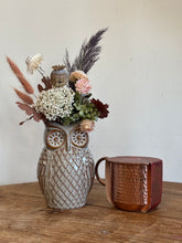 An owl-shaped ceramic vase filled with dried flowers and grasses, placed on a wooden table next to a hammered copper mug against a plain off-white wall.
