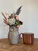 An owl-shaped ceramic vase filled with dried flowers and grasses, placed on a wooden table next to a hammered copper mug against a plain off-white wall.