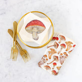 White cocktail napkins printed with red and brown mushrooms, arranged on a white marble surface with gold forks and a gold-rimmed plate.