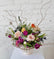 Floral arrangement in pink, purple, peach and white colors in a neutral white ceramic pot. Against a white brick background. 