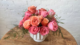 A heart shaped floral arrangement of pink and peach roses with green foliage in a white ribbed ceramic pot, placed on a round wooden table against a white brick wall.