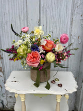 A vibrant, multicolored floral arrangement in a burlap-wrapped vase, displayed on a distressed white wooden table against a weathered gray wood-plank background. 