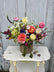 A vibrant, multicolored floral arrangement in a burlap-wrapped vase, displayed on a distressed white wooden table against a weathered gray wood-plank background. 