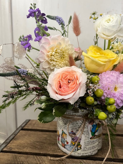 Floral arrangement in soft neutral colors, in a vase decoupaged with pages from a children's book. Arrangement sits atop a wooden crate against a white background.
