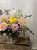 Floral arrangement in soft neutral colors, in a vase decoupaged with pages from a children's book. Arrangement sits atop a wooden crate against a white background.