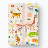 A gift-wrapped rectangular package tied with a pale pink ribbon and bow, covered in a whimsical safari-themed print with an orange tiger, giraffe, crocodile, flamingo, bird, elephant, cat, and gift boxes on an off-white background.