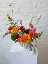 Floral arrangement in bright hues (pink, purple, orange, yellow, greens), set in a neutral ceramic pot, against a white brick wall. 