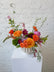 Floral arrangement in bright hues (pink, purple, orange, yellow, greens), set in a neutral ceramic pot, against a white brick wall. 