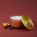 A three-wick candle in a ribbed terracotta glass jar with a gold-toned lid leaning beside it, styled on a dark red background with orange peel and star anise.