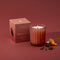 A lit Thymes Pomander Clove scented candle in a ribbed terracotta jar sits next to its dusty brick colored packaging box on a matching background, styled with star anise and a citrus peel slice.