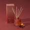 Thymes Pomander Clove reed diffuser set with a frosted terracotta bottle, natural reed sticks, and a matching packaging box on a dusty brick-colored background, styled with orange peel and spices.