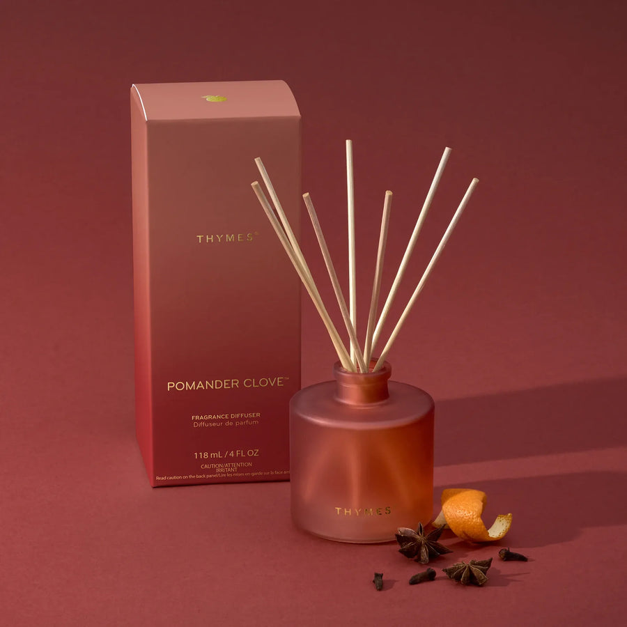 Thymes Pomander Clove reed diffuser set with a frosted terracotta bottle, natural reed sticks, and a matching packaging box on a dusty brick-colored background, styled with orange peel and spices.