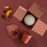 A votive in a ribbed terracotta glass shown in a styled flat lay on a dusty rose background, with an open, patterned box containing a white candle, small decorative spices, and citrus peel.