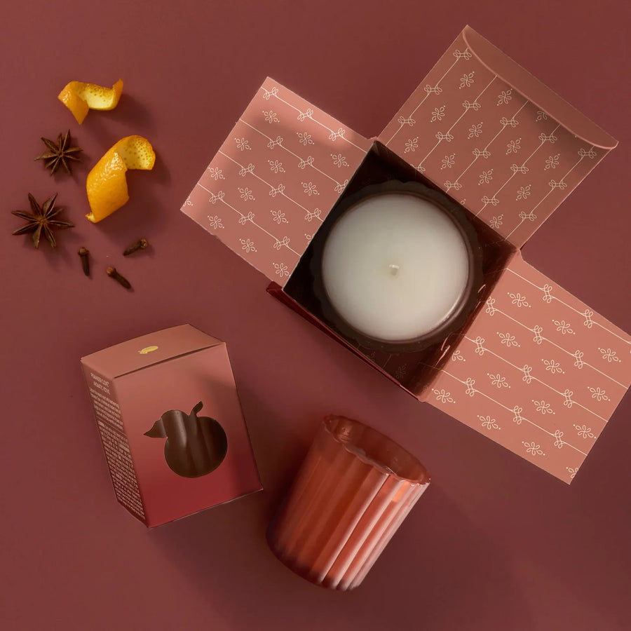 A votive in a ribbed terracotta glass shown in a styled flat lay on a dusty rose background, with an open, patterned box containing a white candle, small decorative spices, and citrus peel.