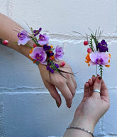 Hands modeling a corsage and boutonniere in purple, pink, and orange hues against a white wall. Corsage and boutonniere are both on wire bases.