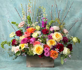 A large, modern-style funeral arrangement in bright, cheerful colors, set in a wooden box against a grey-blue background.
