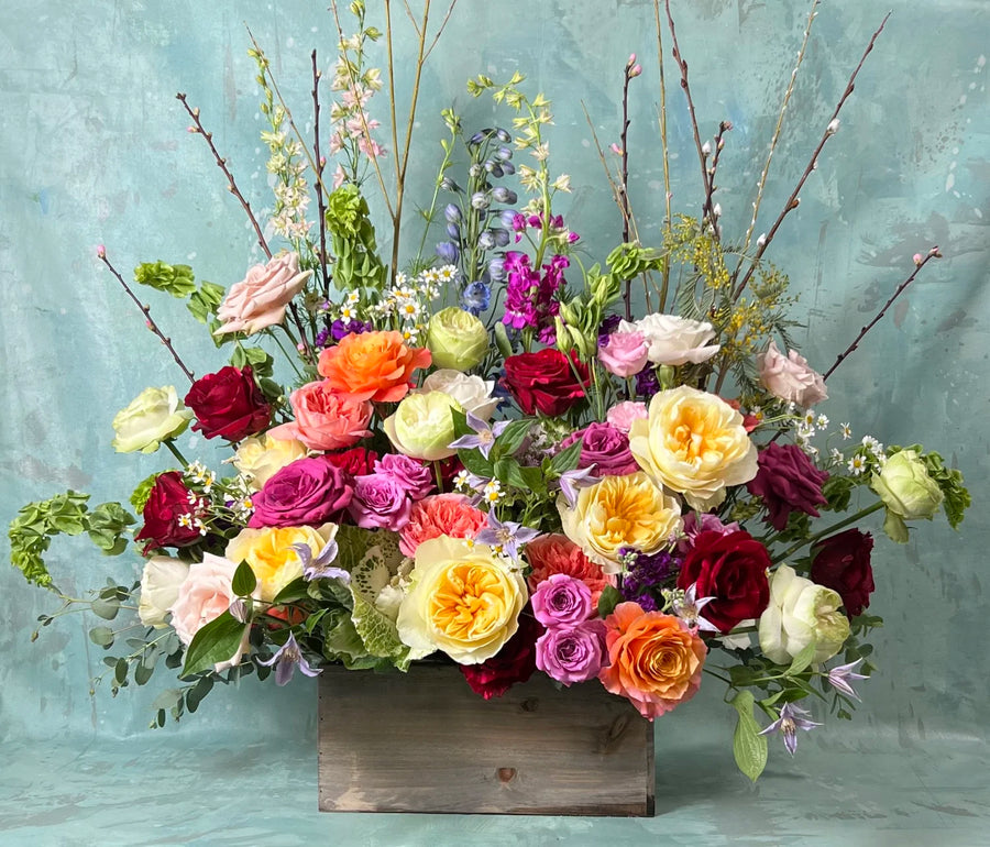 A large, modern-style funeral arrangement in bright, cheerful colors, set in a wooden box against a grey-blue background.