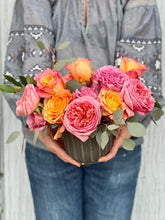 A person in a gray embroidered blouse and blue jeans holding a heart shaped floral arrangement of pink and orange roses with green foliage in a ribbed gray pot against a light gray background.
