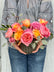 A person in a gray embroidered blouse and blue jeans holding a heart shaped floral arrangement of pink and orange roses with green foliage in a ribbed gray pot against a light gray background.