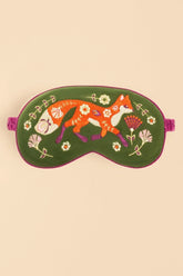 A green embroidered sleep mask featuring a whimsical orange fox, white flowers, and purple botanical details, shown against a solid beige background.