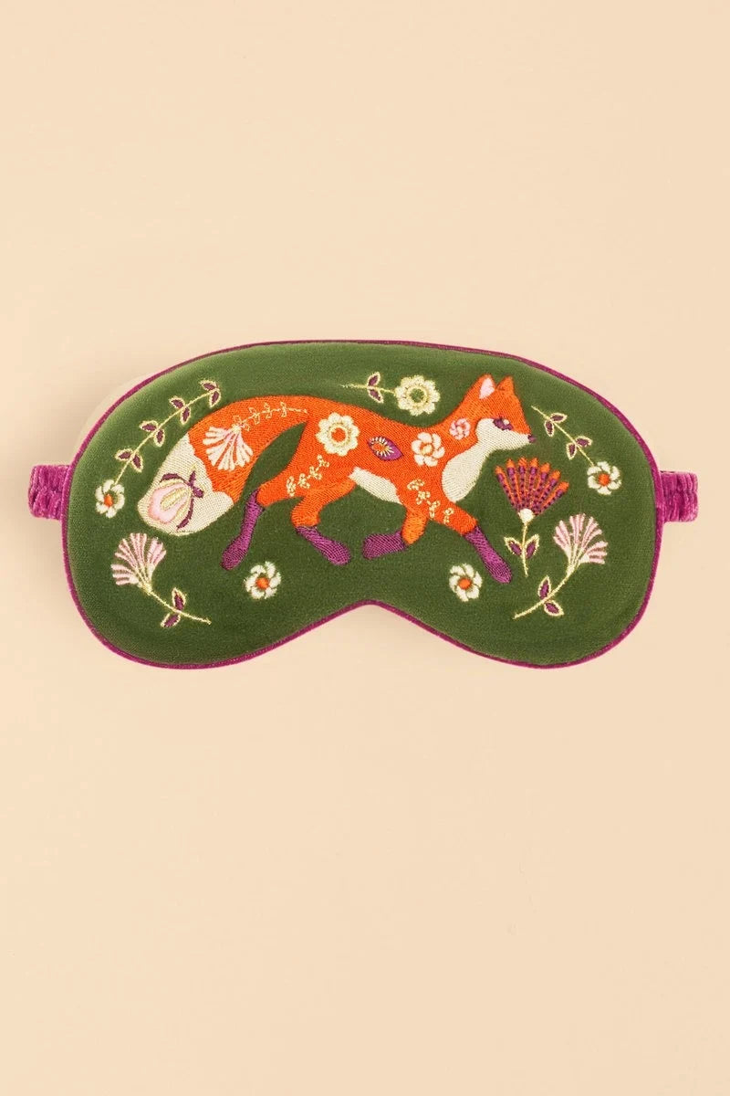 A green embroidered sleep mask featuring a whimsical orange fox, white flowers, and purple botanical details, shown against a solid beige background.