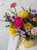 A vibrant floral arrangement in a textured gold vase, featuring bright pink, yellow, and purple blooms with green foliage, set against a white brick wall background.
