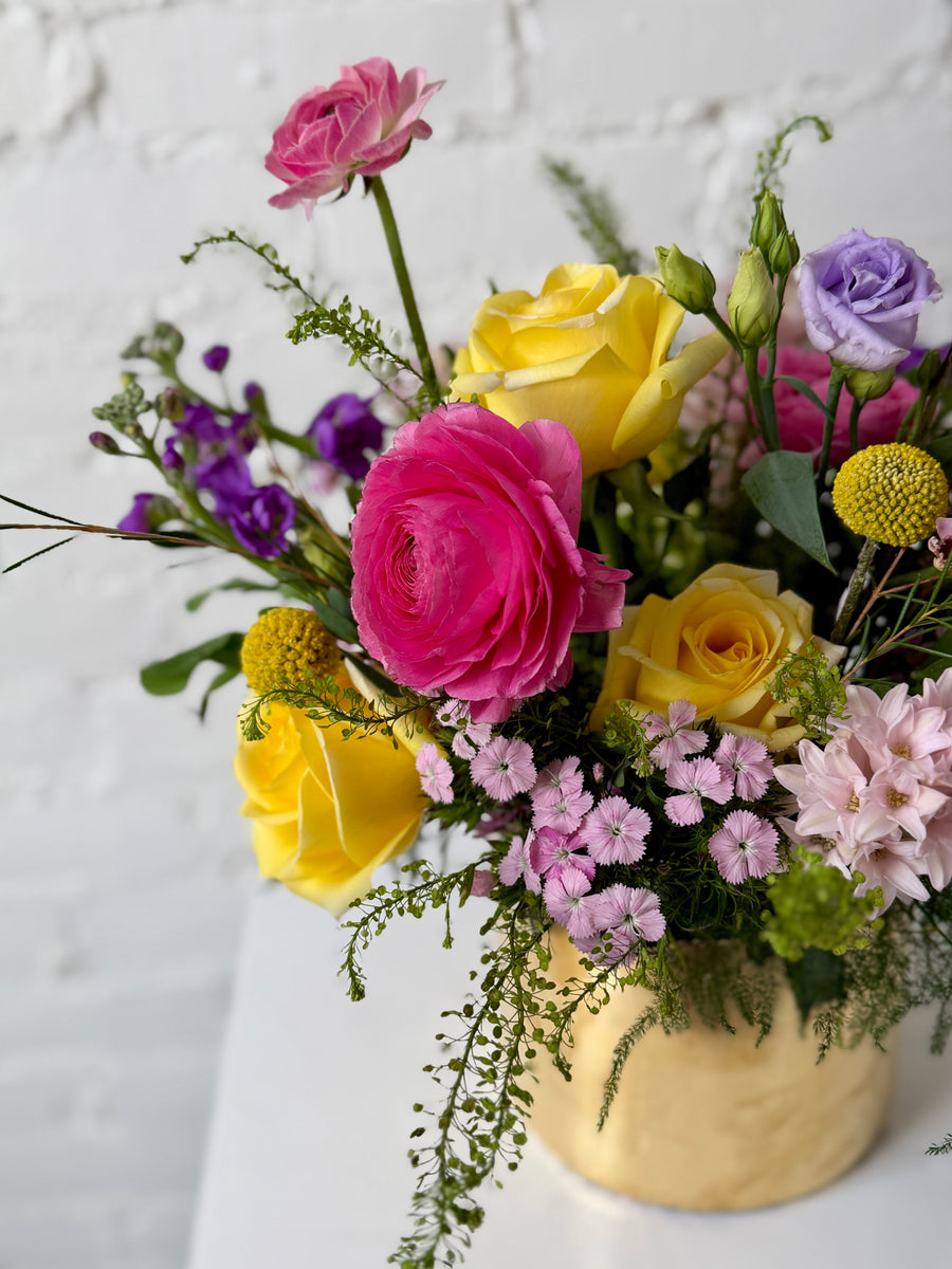 A vibrant floral arrangement in a textured gold vase, featuring bright pink, yellow, and purple blooms with green foliage, set against a white brick wall background.