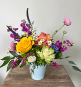 Freeform floral arrangement in bright colors, designed in a plain white ceramic vase. Sat on a wooden table in front of a white background.