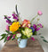 Freeform floral arrangement in bright colors, designed in a plain white ceramic vase. Sat on a wooden table in front of a white background.