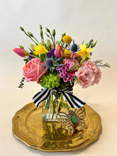 A petit flower arrangement in bright, cheerful colors made in a rectangular glass vase with a black and white striped ribbon around the mouth. Displayed on a gold plate with a metal butterfly brooch, against a light cream background. 