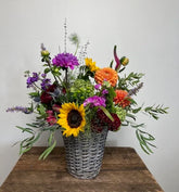 A vibrant mixed flower arrangement in a gray wicker basket, featuring sunflowers, purple and orange flowers, green foliage, and lavender sprigs, displayed on a rustic wooden table against a plain light gray wall.