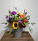 A vibrant mixed flower arrangement in a gray wicker basket, featuring sunflowers, purple and orange flowers, green foliage, and lavender sprigs, displayed on a rustic wooden table against a plain light gray wall.