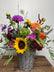 A vibrant wildflower arrangement in a gray woven basket vase, featuring a bright yellow sunflower, purple and orange flowers, green foliage, and burgundy blooms on a rustic wooden table against a plain wall.