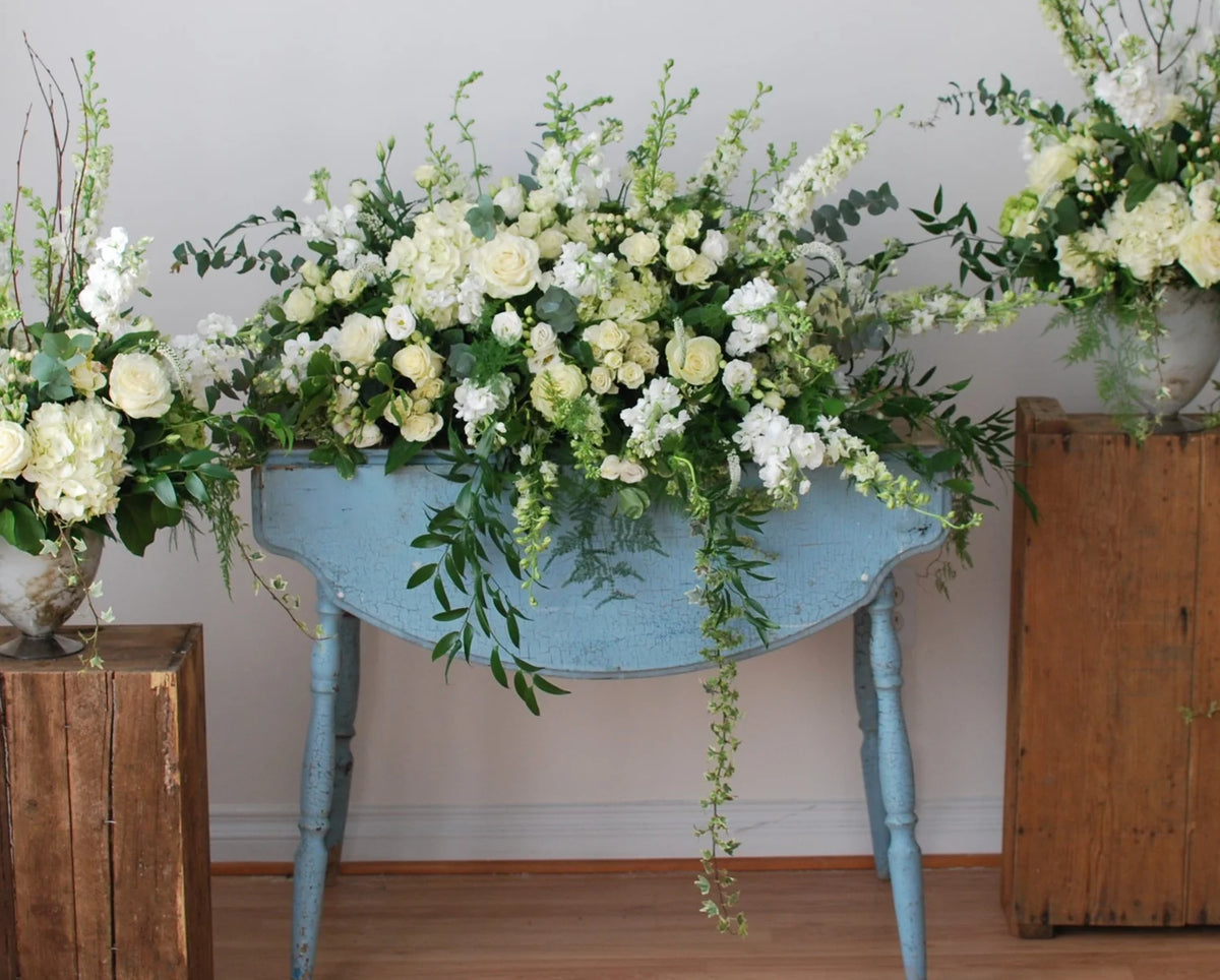 A large casket spray in white and cream roses, hydrangeas, and green foliage, displayed on a distressed light blue console table. The table is flanked by rustic wooden crates holding additional floral arrangements against a plain off-white wall and a light wood floor.